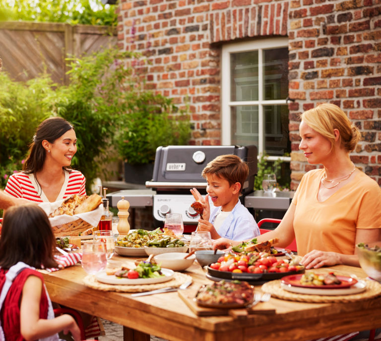 Lifestyle image of a family enjoying food together outdoors with the Weber Spirit E-310 gas barbecue in soft focus in the background, creating a relaxed and premium summer entertaining scene.