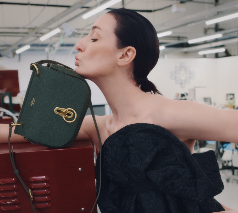 Smiling woman in a white bodysuit waving while lying on a factory trolley in the Mulberry Somerset factory, holding a tan Mulberry handbag and wearing black loafers