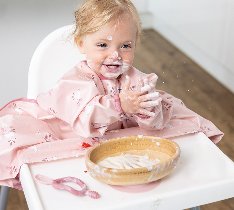 Image shows a toddler in a highchair, wearing a Bibado curious cottontail coverall, with a Bibado Bamboo suction bowl on the highchair.