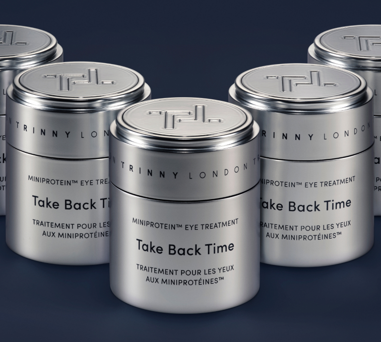An image of Trinny London's Take Back Time miniprotein™ eye treatment or "traitement pour les yeux aux miniproteines™". Multiple silver pots arranged in a V-formation against a navy blue background.