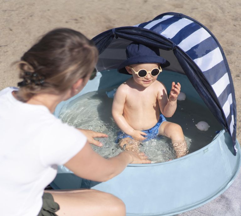 The scene is set alongside a swimming pool in the sun, a pop-up anti tent is set up in the foreground aside from the swimming pool next to some sun loungers.