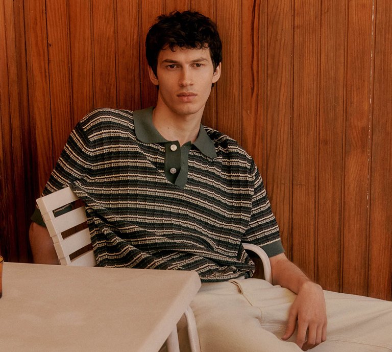 Men’s summer fashion banner featuring a model in a striped polo shirt and light trousers, styled in a warm-toned setting, highlighting lightweight layers and relaxed, everyday dressing.